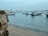 Several small fishing boats are anchored near the shore with a distant view of modern high-rise buildings on the horizon. The water is calm, reflecting the clear blue sky, and the shoreline is composed of fine sand and a rocky foreground.