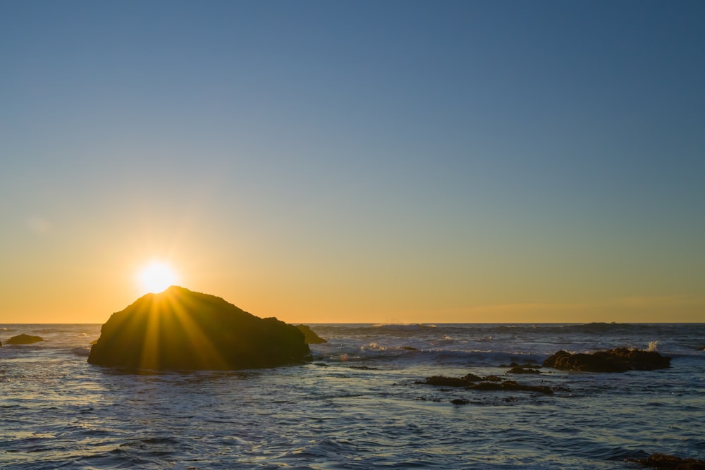 the sun is setting over the ocean with a rock in the foreground