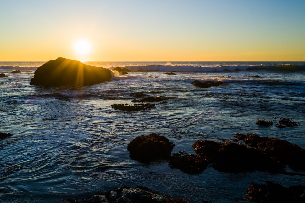 the sun is setting over the ocean with rocks in the foreground