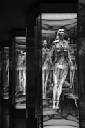 A series of humanoid robotic figures, encased in glass, with intricate mechanical details. The reflections create a tunnel effect, enhancing a futuristic and technological atmosphere.