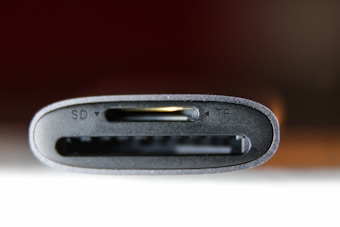 A close-up view of a card reader with slots labeled SD and TF, showing the edges of the slots in a dark gray finish. The background is blurred with warm, dark tones.