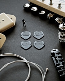 A close-up view of various guitar components, including a set of four black guitar picks, a figure of a person playing guitar, a vacuum tube, and other hardware parts like strings, tuning pegs, and a pickup cover, all arranged on a dark surface.