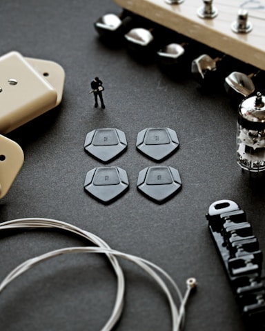 A close-up view of various guitar components, including a set of four black guitar picks, a figure of a person playing guitar, a vacuum tube, and other hardware parts like strings, tuning pegs, and a pickup cover, all arranged on a dark surface.
