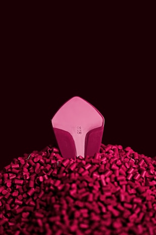 A geometric object with a pink reflective surface protrudes from a mound of small cylindrical pieces. The background is a deep, rich burgundy color, giving the scene a stylish and modern look.