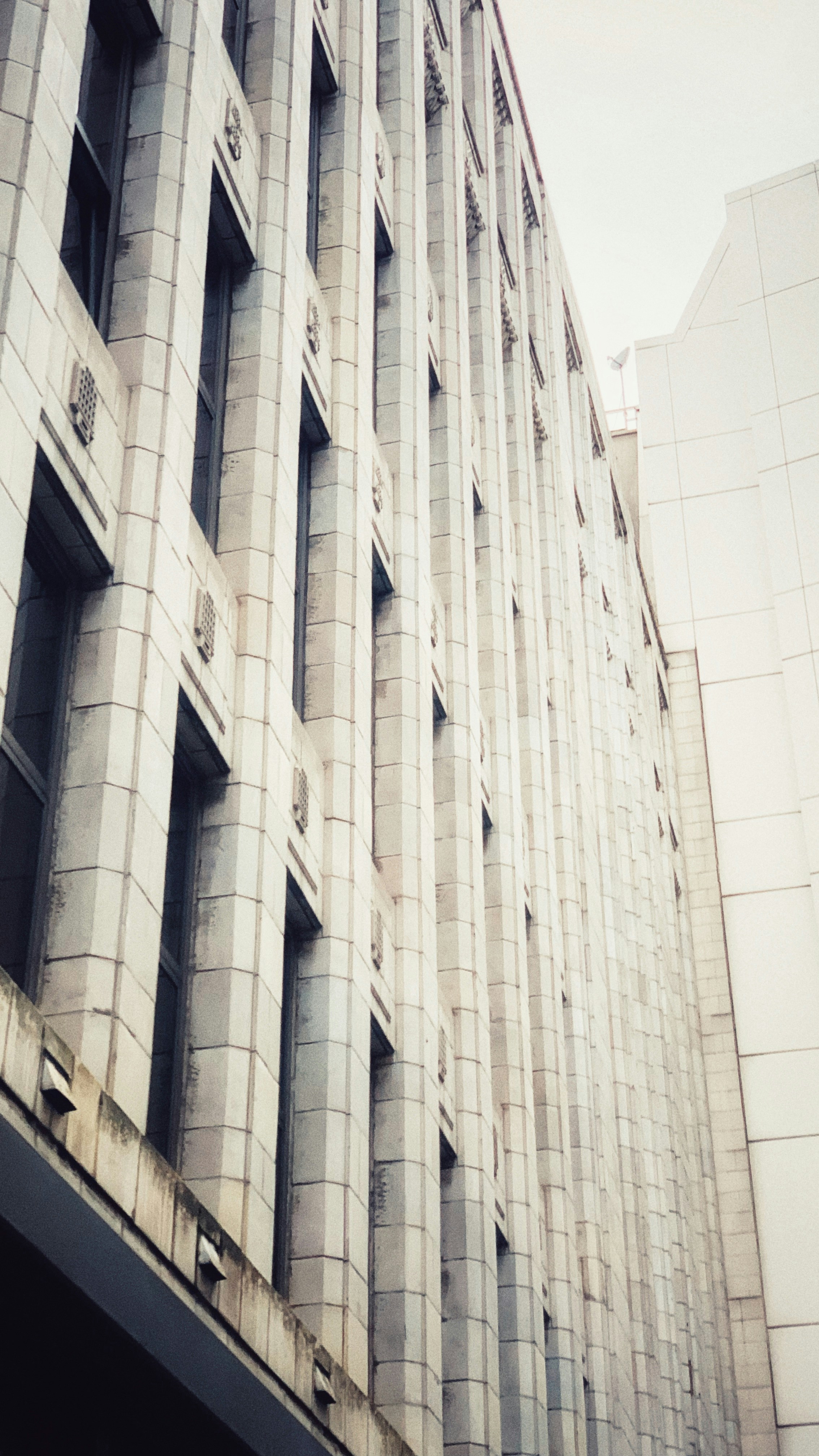 An architectural view of a tall, multi-story building with a modern design characterized by tall, vertical windows and a geometric pattern on the facade. The building has a sleek appearance with a mostly monochromatic color scheme.