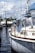 A sailboat is docked in a marina with its sails neatly wrapped and secured. The boat features a classic design with a blue and white color scheme. Reflections in the calm water mirror the vessel, while other boats are visible in the background. The scene is set under a partly cloudy sky, suggesting pleasant weather conditions.