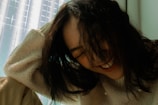 A person with long dark hair is captured in a moment of laughter, wearing a cream-colored fuzzy sweater. The background features a softly lit space, with a grid-like pattern on a translucent surface.