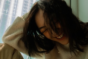A person with long dark hair is captured in a moment of laughter, wearing a cream-colored fuzzy sweater. The background features a softly lit space, with a grid-like pattern on a translucent surface.