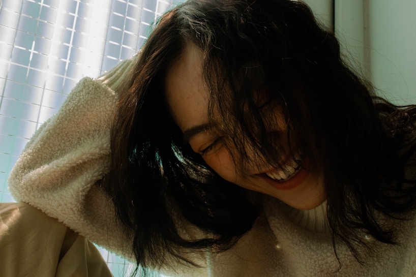 A person with long dark hair is captured in a moment of laughter, wearing a cream-colored fuzzy sweater. The background features a softly lit space, with a grid-like pattern on a translucent surface.
