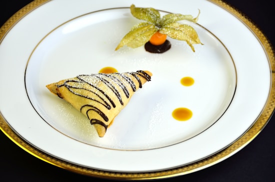 A meticulously presented dessert featuring a triangular pastry with chocolate drizzle and a dusting of powdered sugar. It is served on a white plate with a decorative gold rim. Accompanying the pastry is a small orange sphere, possibly a fruit or sauce, garnished with a leafy husk, and additional sauce droplets adding a touch of elegance.