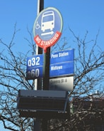 A public transportation sign is mounted on a pole, indicating bus routes Q32 and Q60 with destinations such as Penn Station and Midtown. The sign has blue and white colors with a prominent circular 'No Standing' notice with a red border and a bus icon. The background includes leafless branches against a clear blue sky.