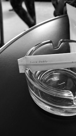 A clear glass ashtray sits on a dark surface. Resting in the ashtray is a paper packet labeled 'Sucre Daddy'. In the background, part of a person's leg is visible, slightly out of focus.