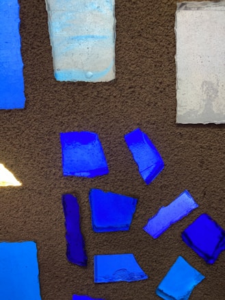 A collection of variously shaped blue and light blue glass fragments are set against a textured brown background. The pieces have rough edges and display different shades of blue, ranging from deep cobalt to lighter azure. The arrangement of the glass forms a scattered pattern, with the light reflecting off their surfaces, creating subtle highlights.