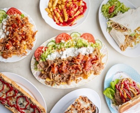 An assortment of various fast food dishes is arranged on a light-colored table. The selection includes plates of doner kebab with vegetables and sauces, fries drizzled with ketchup, a wrap filled with vegetables and meat, a loaded sandwich topped with ketchup, and a side dish likely consisting of a baked item with melted cheese. The presentation is colorful and appetizing.