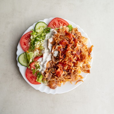 A white plate filled with a serving of doner kebab meat, fresh sliced tomatoes, cucumber, and lettuce, accompanied by creamy white and red sauces.