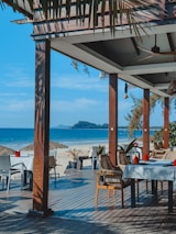 A beachfront dining area with tables and chairs set under a wooden pergola. The setup is on a wooden deck overlooking a sandy beach and a calm blue ocean. There are thatched umbrellas on the beach and lush greenery in the surroundings. The scene is sunlit, suggesting a warm and inviting atmosphere.