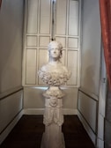 A detailed bust sculpture of a person on a pedestal, located in a room with elegant decor. The sculpture has intricate carvings and displays a calm expression. The pedestal is adorned with floral and decorative motifs. In the background, there is a set of paneled doors that add to the classic and sophisticated setting.