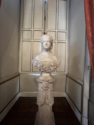 A detailed bust sculpture of a person on a pedestal, located in a room with elegant decor. The sculpture has intricate carvings and displays a calm expression. The pedestal is adorned with floral and decorative motifs. In the background, there is a set of paneled doors that add to the classic and sophisticated setting.