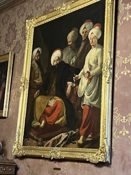A large, ornate painting in a gilt frame hangs on a wall with intricate wallpaper. The painting depicts five men wearing traditional Middle Eastern or South Asian clothing with turbans. The central figure is seated while the others stand around, and they all appear to be engaged in a serious discussion.