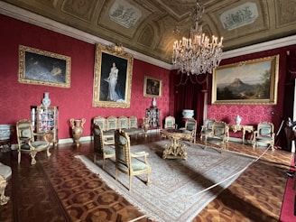 An opulent, classical sitting room features ornate furniture with gilded accents and upholstered chairs arranged around a large, intricate carpet. The room is decorated with large paintings in gilded frames, elegant vases, and a grand chandelier. The walls are covered in a deep red patterned wallpaper, enhancing the luxurious ambiance.