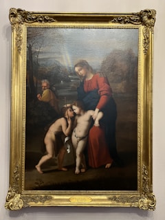 A classical painting depicts a serene scene with a woman in a red robe and two young children, one holding a cross, standing in a pastoral landscape. An elderly man watches over them in the background, and a faint rainbow arcs above the horizon.