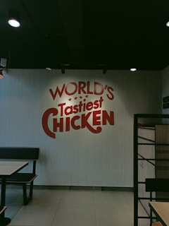A restaurant interior with a wall featuring red text that reads 'WORLD'S Tastiest CHICKEN'. The space has a simple and modern design with black booth seating and tables. The lighting is soft, creating a relaxed atmosphere.