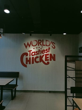 A restaurant interior with a wall featuring red text that reads 'WORLD'S Tastiest CHICKEN'. The space has a simple and modern design with black booth seating and tables. The lighting is soft, creating a relaxed atmosphere.
