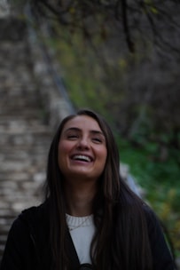 A person with long dark hair is laughing joyfully against a blurred background of stone steps and greenery. The overall atmosphere suggests a moment of happiness in an outdoor setting.