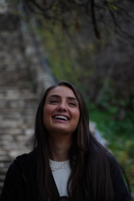 A person with long dark hair is laughing joyfully against a blurred background of stone steps and greenery. The overall atmosphere suggests a moment of happiness in an outdoor setting.
