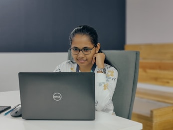A person with glasses is sitting at a desk, focused on a laptop with a Dell logo. The individual is wearing a light-patterned top and a wristwatch, and appears to be in a modern office environment with wooden seating in the background.