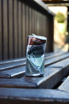 A can of Starbucks Doubleshot Espresso stands on a wooden bench. The can is slightly crushed at the top and features a combination of brown and green colors with the Starbucks logo prominently displayed. The image focuses on the can, with the wooden background and seating creating a rustic and casual atmosphere.