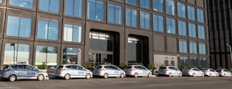 A line of several NYPD police cars parked in front of a modern building with large reflective windows. The architecture features an industrial design with repetitive rectangular patterns.