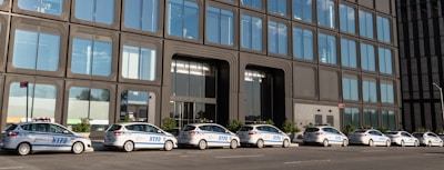 A line of several NYPD police cars parked in front of a modern building with large reflective windows. The architecture features an industrial design with repetitive rectangular patterns.