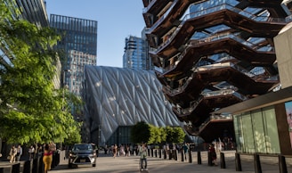 A modern urban scene featuring contemporary architecture. The left side shows a large, uniquely textured building with diagonal lines and a metallic appearance. On the right, a distinctive structure with a honeycomb-like design and reflective surfaces is visible. In the foreground, there are people walking and a car parked on the street. Green trees and glass skyscrapers add to the vibrant urban environment.