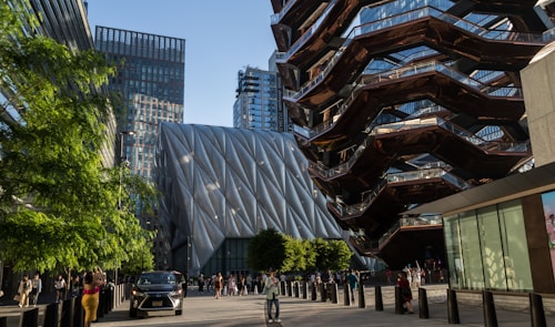 A modern urban scene featuring contemporary architecture. The left side shows a large, uniquely textured building with diagonal lines and a metallic appearance. On the right, a distinctive structure with a honeycomb-like design and reflective surfaces is visible. In the foreground, there are people walking and a car parked on the street. Green trees and glass skyscrapers add to the vibrant urban environment.