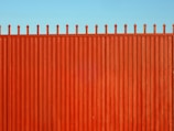 A tall, vertical red fence with evenly spaced slats and pointed tops stands against a clear blue sky.