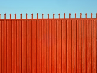 A tall, vertical red fence with evenly spaced slats and pointed tops stands against a clear blue sky.