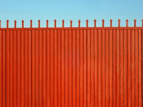 A tall, vertical red fence with evenly spaced slats and pointed tops stands against a clear blue sky.