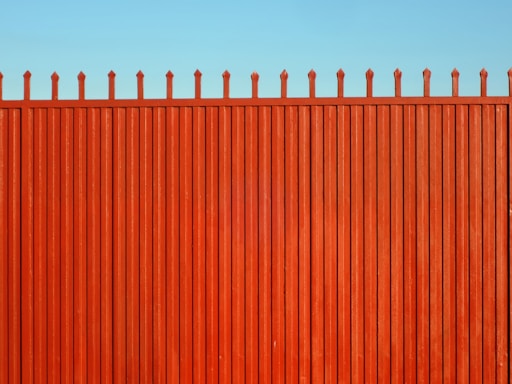 A tall, vertical red fence with evenly spaced slats and pointed tops stands against a clear blue sky.