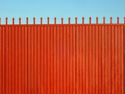 A tall, vertical red fence with evenly spaced slats and pointed tops stands against a clear blue sky.