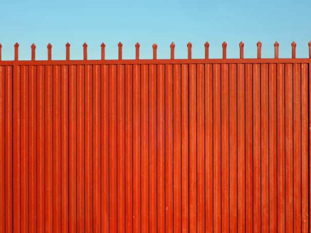 A tall, vertical red fence with evenly spaced slats and pointed tops stands against a clear blue sky.