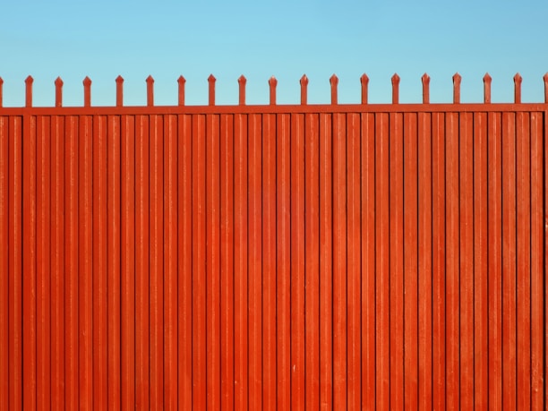 A tall, vertical red fence with evenly spaced slats and pointed tops stands against a clear blue sky.