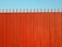 A tall, vertical red fence with evenly spaced slats and pointed tops stands against a clear blue sky.