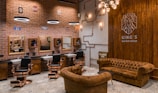 A stylish barbershop interior featuring brick walls adorned with vintage signs and a prominent Route 66 theme. There are multiple barber stations with mirrors and black cushioned chairs featuring copper accents. The seating area includes brown tufted leather sofas and chairs. Modern lighting fixtures and exposed copper piping add an industrial touch to the cozy ambiance.