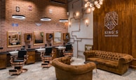 A stylish barbershop interior featuring brick walls adorned with vintage signs and a prominent Route 66 theme. There are multiple barber stations with mirrors and black cushioned chairs featuring copper accents. The seating area includes brown tufted leather sofas and chairs. Modern lighting fixtures and exposed copper piping add an industrial touch to the cozy ambiance.