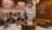A stylish barbershop interior featuring brick walls adorned with vintage signs and a prominent Route 66 theme. There are multiple barber stations with mirrors and black cushioned chairs featuring copper accents. The seating area includes brown tufted leather sofas and chairs. Modern lighting fixtures and exposed copper piping add an industrial touch to the cozy ambiance.