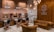 A stylish barbershop interior featuring brick walls adorned with vintage signs and a prominent Route 66 theme. There are multiple barber stations with mirrors and black cushioned chairs featuring copper accents. The seating area includes brown tufted leather sofas and chairs. Modern lighting fixtures and exposed copper piping add an industrial touch to the cozy ambiance.
