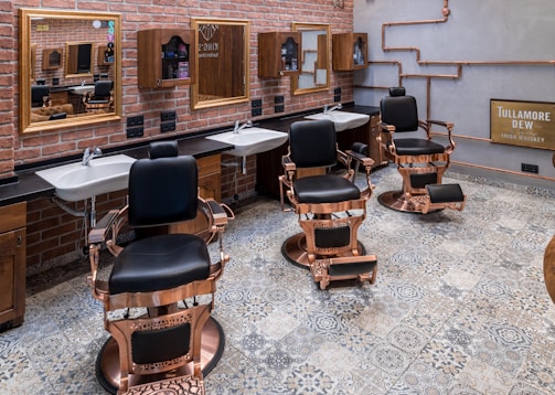 A stylish barbershop interior with three ornate barber chairs featuring black cushions and bronze detailing. The room has a rustic brick wall with two mirrors above white sinks and wooden cabinets. The floor is covered with patterned tiles, and copper pipes are visible on the side wall next to a Tullamore Dew whiskey sign.