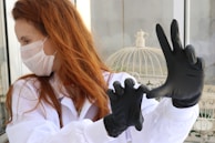 A person with red hair wearing a white lab coat and a white mask is adjusting black gloves on their hands. In the background, there is a white birdcage with a decorative element inside a room with glass windows.