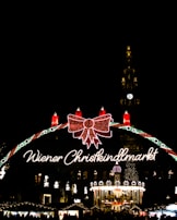 A festive Christmas market scene illuminated at night with a large decorative sign reading 'Wiener Christkindlmarkt'. The sign features a large red bow and several lit candles. Below, a carousel is brightly lit and surrounded by decorated stalls. In the background, a tall clock tower is lit up, with the clock face clearly visible.